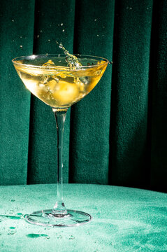 Vibrant yellow cocktail with ice splashing in a modern coupe glass against a luxurious emerald green velvet background, creating a dynamic bar or party scene