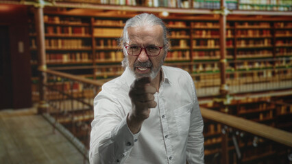 Man points finger at camera in library building among bookshelves with clenched jaw and raised hand  anger confrontation authority. © Krakenimages.com