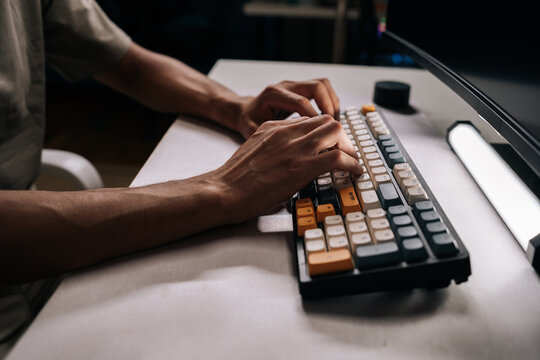 Side view of male hands typing on mechanical keyboard with colorful keycaps in dimly lit office, suggesting focused work or late-night coding session. Concept of modern technology.
