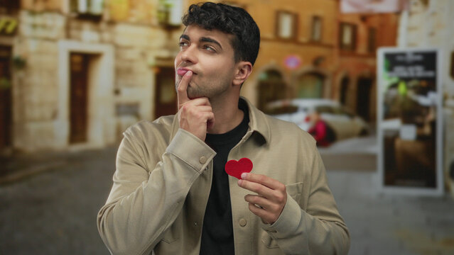 Man holds heart while pondering on street with young hispanic thinking male feelings about love in urban setting.