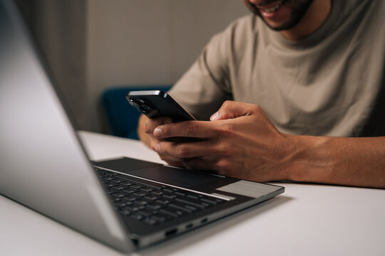 Smiling young businessman working late at night in home office, using smartphone and laptop to manage business tasks and meet deadlines with focused concentration and professionalism.