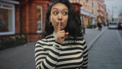 Young hispanic woman with finger to lips for silence on a city street wearing striped sweater and hoop earring  quiet secrecy. © Krakenimages.com
