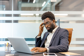 African american businessman sitting at office desk, experiencing stress and thinking deeply while working on his laptop, focusing on complex tasks and solving problems