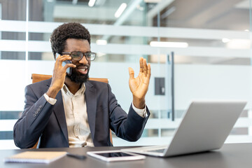 African american businessman managing a phone call and gesturing while working at his desk with a laptop and tablet in a modern corporate office environment