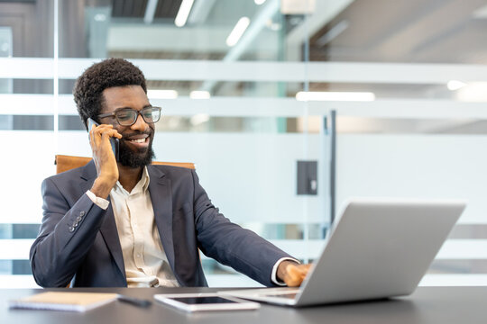 Smiling young african american businessman sitting at office desk, having a phone conversation, and using a laptop for remote work and corporate communication - Powered by Adobe