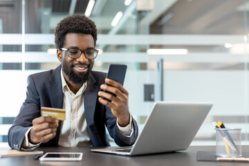 Smiling african american man wearing glasses making a secure online payment, holding a credit card and smartphone, with a laptop on a desk in a modern office