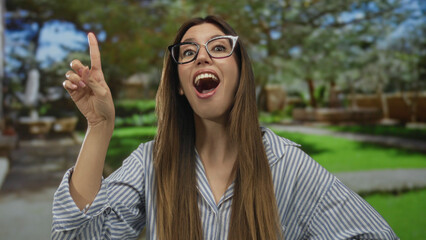 Young hispanic woman wearing glasses and striped shirt finger pointing upward in lush forest; curiosity.