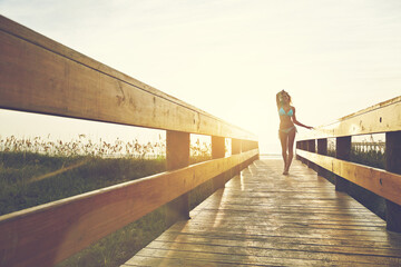Obraz premium Happy woman, bridge or boardwalk at beach with sunset, low angle space and vacation for summer travel. Adventure, portrait or person in bikini for coastal holiday, sunshine and walk on wood pathway