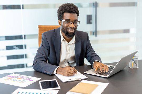African american businessman working at his office desk, typing on a laptop, smiling at the camera, and writing notes on a clipboard with documents and a tablet around him
