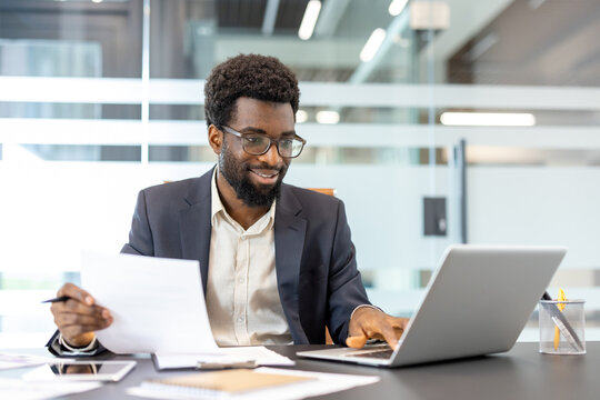 African american businessman wearing glasses smiling and working proactively at an office desk, analyzing documents and typing on a laptop with a modern background