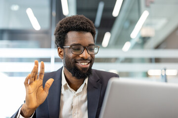 Smiling african american businessman communicating remotely during a professional video call, sitting at his office desk and gesturing to a laptop screen