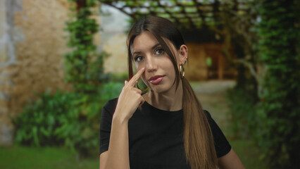 Fototapeta premium Woman with long brown hair wearing gold hoop earrings touches her eye with a finger at building entrance; focus calm.