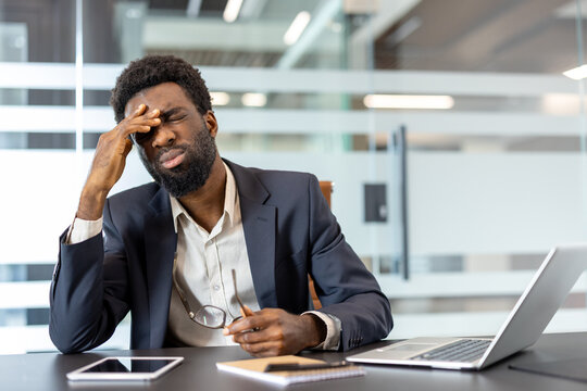 Unhappy young african american businessman experiencing headache and stress while sitting at desk in modern office, feeling overwhelmed and fatigued from professional workload - Powered by Adobe