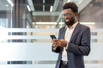 Young businessman in a suit standing in a modern office, smiling while using his mobile phone, connecting with technology and digital communication © Liubomir