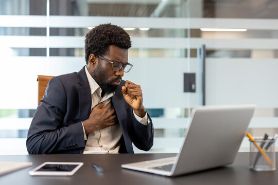 African american businessman sitting at his desk in a modern office, feeling unwell and coughing into his fist, experiencing chest pain and discomfort while working