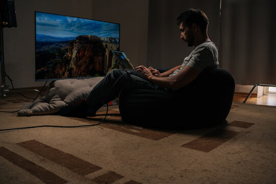 Video editor sitting on beanbag chair using laptop for editing video or photo content, with big screen tv showing footage in background. Concept of modern technology.