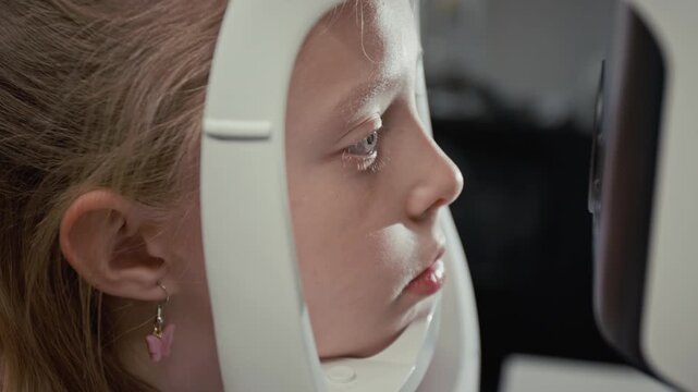 Side view close up shot of young girl looking into diagnostic machine during eye exam in clinic with focus on blue eyes