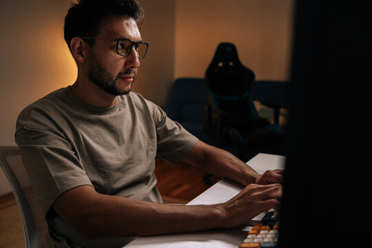 Focused programmer freelancer working on desktop computer late at night, illuminated by soft glow of a lamp and blue light of mouse and keyboard. Concept of modern technology.