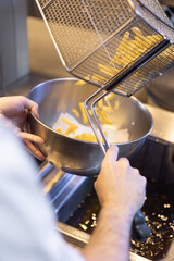 Freshly fried French fries being poured from a metal fryer basket into a stainless steel bowl inside a professional kitchen. The warm light, the motion of the falling fries, and the industrial