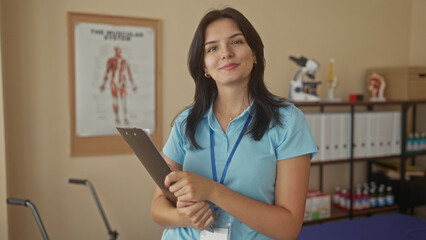 Young hispanic woman holds clipboard and touches hair in building near anatomy poster and...