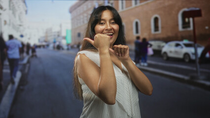 Young latina brunette woman smiling with thumb pointing sideways on a city street by brick building; playful confidence.