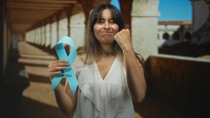 Young latina brunette woman holding blue awareness ribbon and fist raised in old building corridor;...