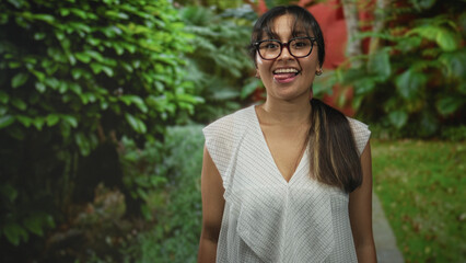 Young latina woman with glasses and ponytail, wearing a white blouse, sticking out her tongue and raising both hands in a green park path; playful mischief.