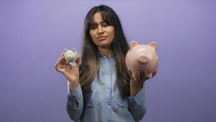 Young woman holding two piggy banks and comparing a small white and large pink bank in a purple...