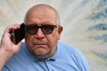 An elderly man wearing glasses and a blue polo shirt and glasses holds a phone to his ear.