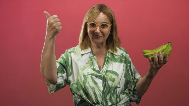 Middle age woman with green leaf shirt holding bananas and giving thumbs up in studio; cheerful healthy living. - Powered by Adobe