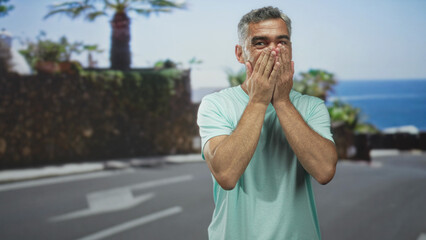 Man with hands covering mouth on a seaside street by the ocean and palm trees, wearing a green tshirt showing an expressive face; surprise wonder.