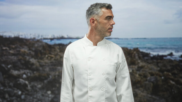 Man in chef jacket looking right on rocky coast with ocean and waves behind, subtle head turn and open mouth gesture; contemplation. - Powered by Adobe