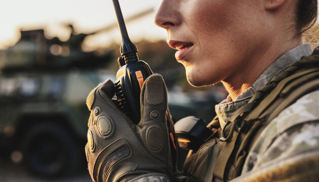 Close-up of a female soldier in camouflage uniform communicating via walkie-talkie with a military vehicle in the background.