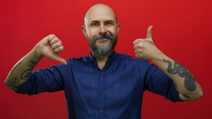 Bald man with beard showing approval and disapproval gestures against red background, wearing blue...