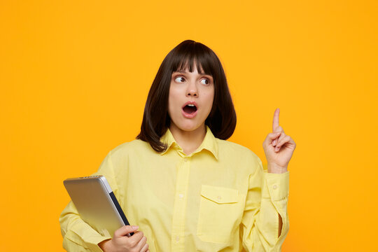 A surprised young woman in a yellow shirt holds a tablet, pointing up as she reacts to a surprising idea. The bold orange backdrop adds energy and focus to the scene. - Powered by Adobe