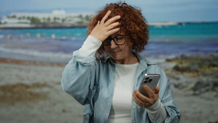 Woman on beach holding smartphone appears frustrated while enjoying seaside view, dressed casually and standing on sandy coastline with blurred background of water and sky.