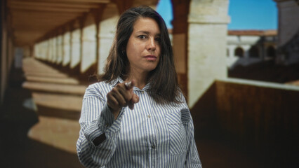 Woman brunette points finger then taps temple in building colonnade with archway corridor, steady gaze toward camera; thoughtful reflection.