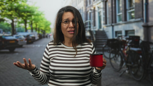 Woman holding red mug, touching hair and gesturing on a city street with bicycles and cobblestone townhouses; thoughtful reflection. - Powered by Adobe