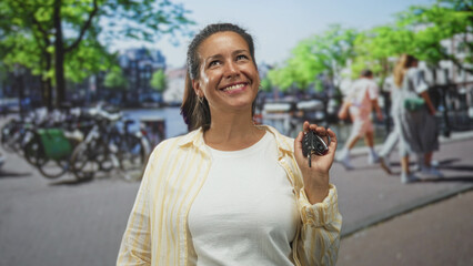 Woman holding car keys and smiling on street canal with parked bicycles, wearing striped yellow shirt over white tee, facing camera and posing; joy.