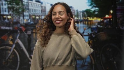 Fototapeta premium Woman smiling, hand to hair, face visible, wearing sweatshirt, standing by bicycle along canal on street; joyful confidence.