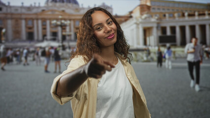 Obraz premium Young african american woman in a beige shirt points finger to camera in st peters square outside the vatican among tourists and colonnades; confidence travel invitation.