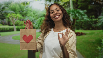 Woman holds paper bag with red heart by the handle in a green park while presenting it with palm up; gift giving kindness joy.