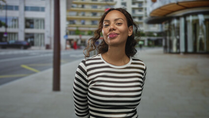 Young woman smiling with hands behind her back on a city street, striped shirt and exposed neck and face; confidence warmth optimism.