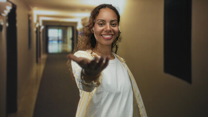 Woman smiling extends her hand toward camera in hotel building corridor wearing white tee and open...