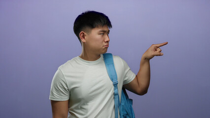 Young man wearing white shirt with blue backpack excitedly pointing at something over isolated purple background