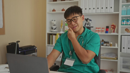 Young chinese male doctor in clinic room wearing green scrubs, using a laptop and phone with medical equipment in the background, focused on communication.