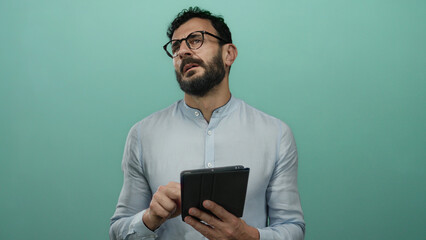 Hispanic man with beard and glasses using a tablet against a green background, wearing a blue shirt and appearing focused and thoughtful.