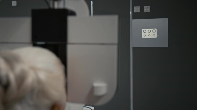 Back view shot of white haired senior woman looking into automated phoropter during eye exam in clinic with focus flowing from patient to eye chart projected on wall, copy space