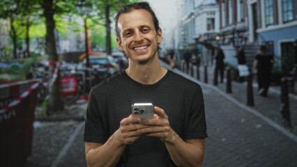 Man holding smartphone on a cobbled city street smiling while looking at screen and holding phone with both hands; casual joy.