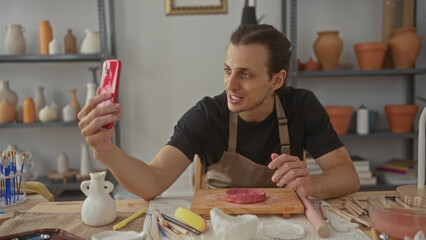 Man holds red smartphone and molds pink clay with his hands at a pottery worktable in studio; craftsmanship focus.